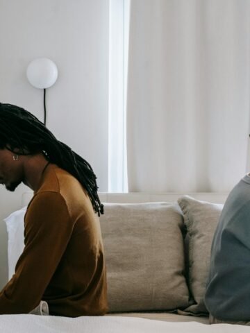 A couple sits back-to-back on a bed, looking upset and distant from each other, suggesting tension or conflict in their relationship.